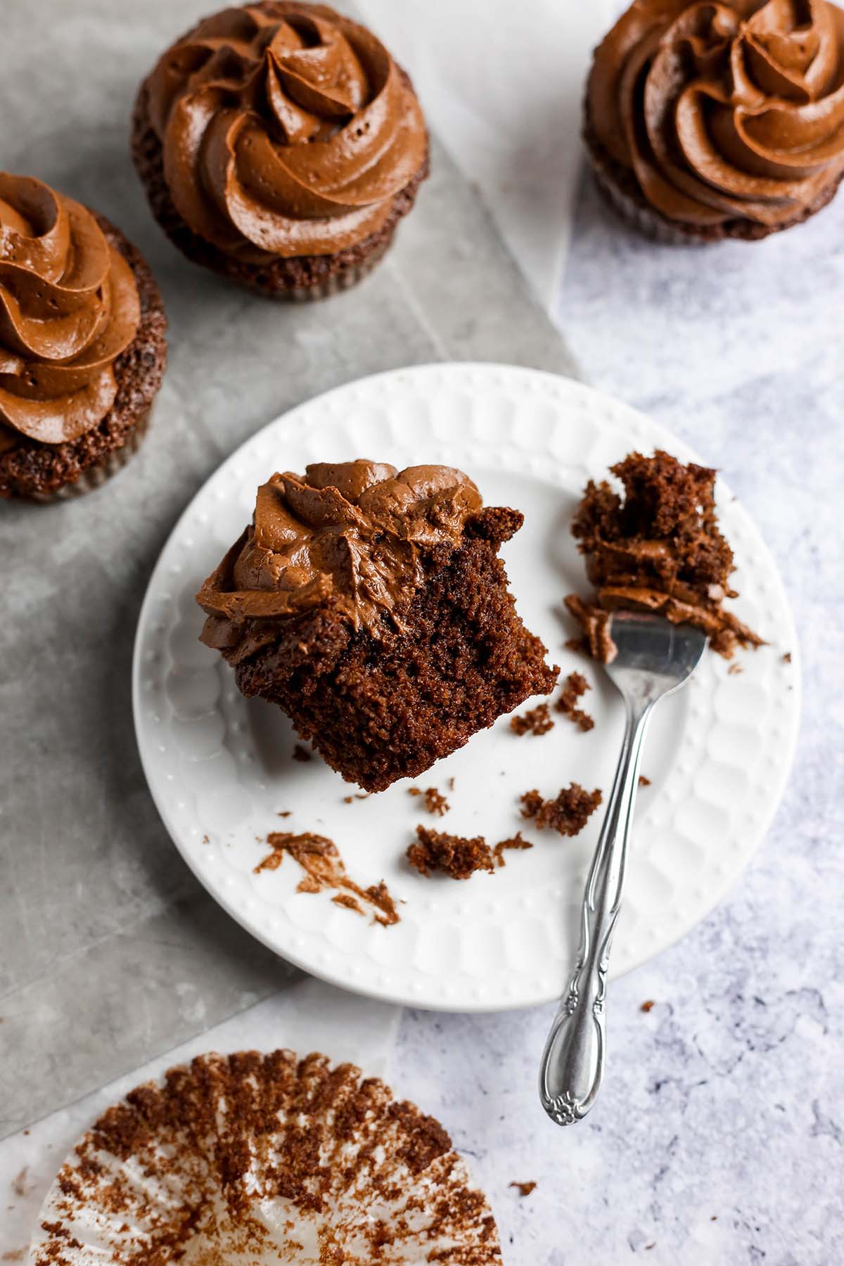 half of a chocolate cupcake with chocolate frosting on a small white plate. A knife is next to the cupcake that is cut in half.