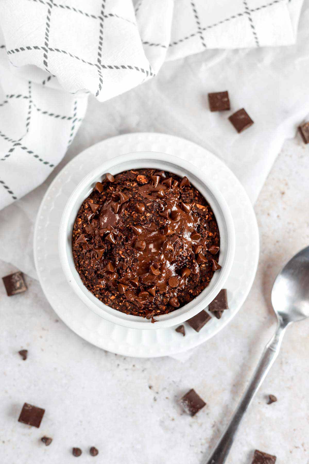 a ramekin of chocolate baked oats on a small dessert plate, with a spoon next to the plate. There is melted chocolate on top of the oats.