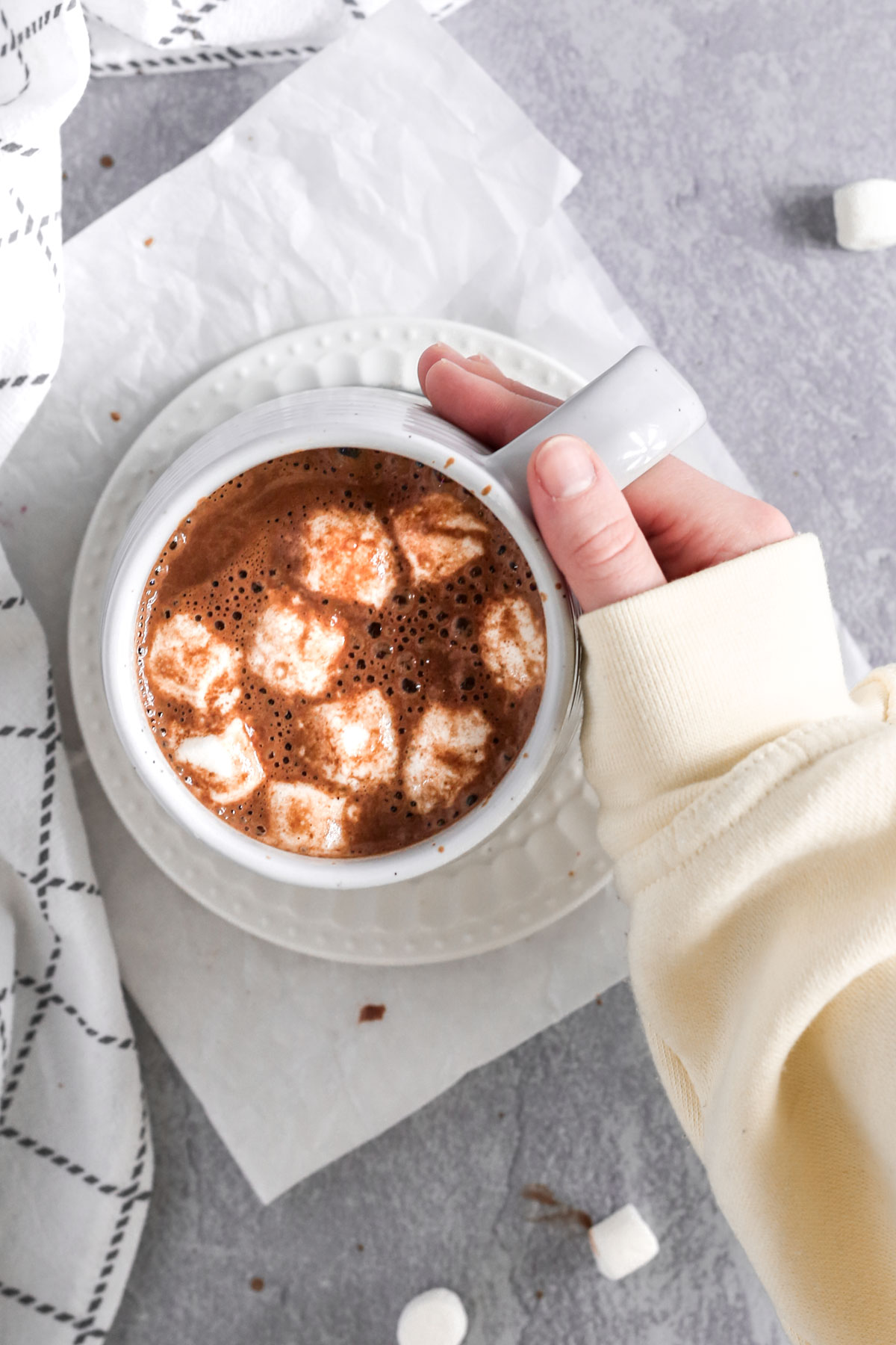 a hand placed around the warm mug of hot cocoa. There are marshmallows in the mug.