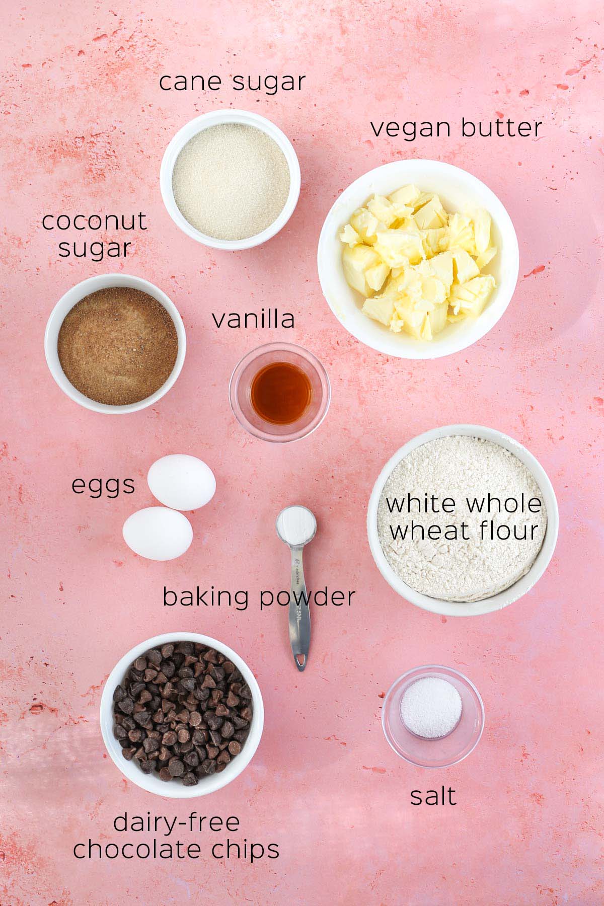 all of the ingredients to make dairy free chocolate chip cookies, set out in small dishes on a pink surface.