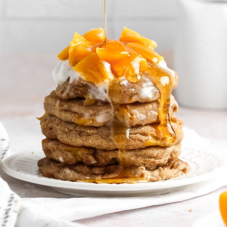 a stack of peach cobbler pancakes on a plate with fresh peaches and maple syrup on top.