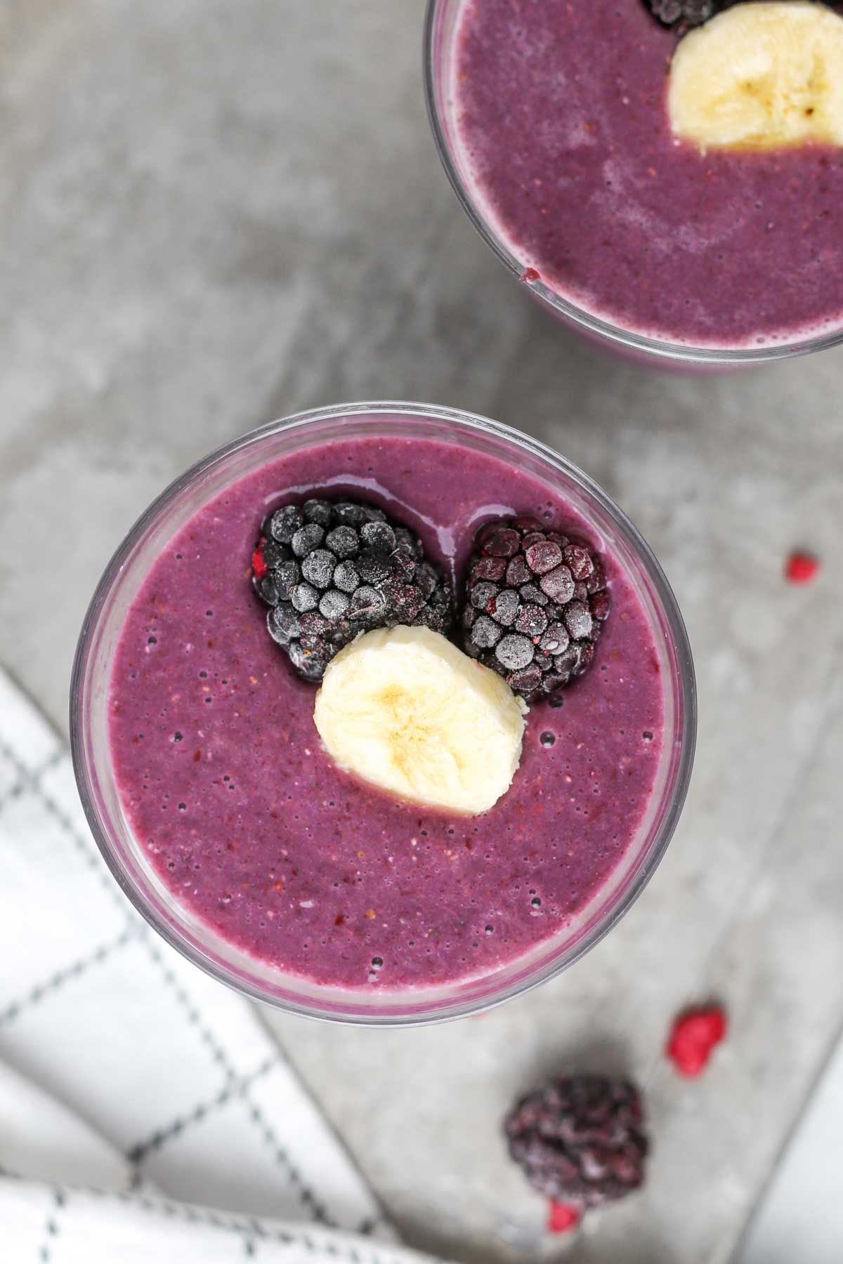 two glasses of purple blackberry banana smoothies with frozen blackberries and fresh bananas on top.