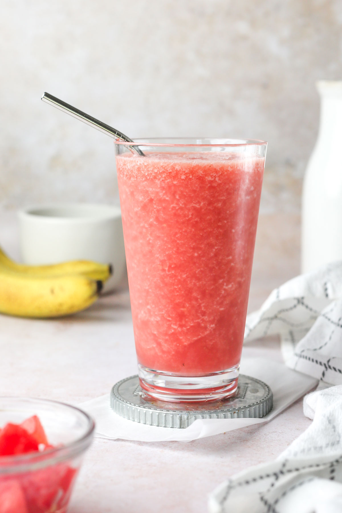 a clear glass of pink watermelon banana smoothie, surrounded by bananas and watermelon.
