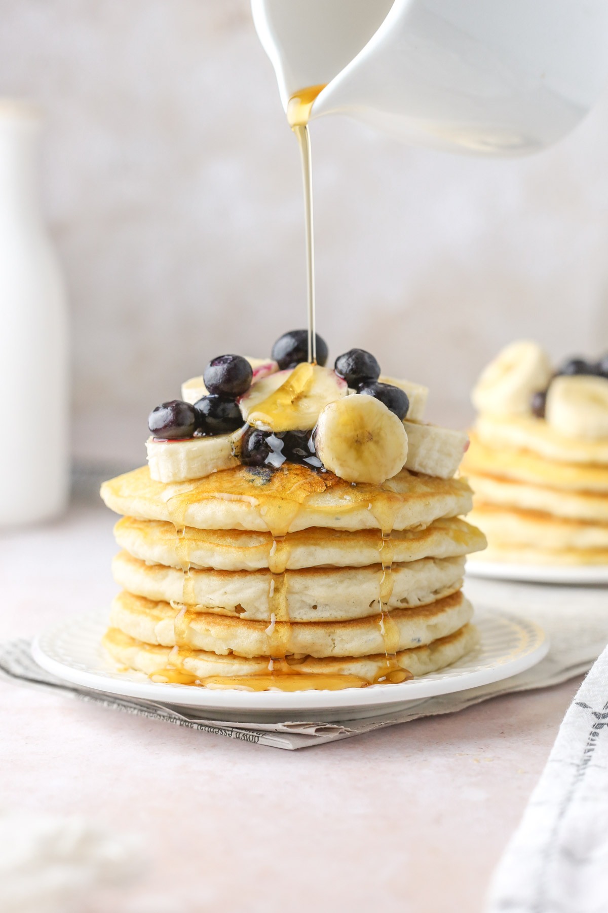 a stack of fluffy oat milk pancakes shown from the side, topped with fresh banana and blueberries, with a drizzle of maple syrup being poured over top.