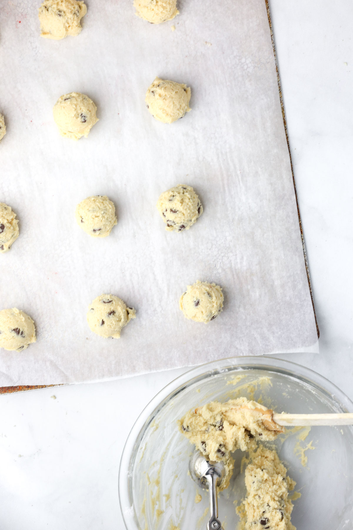 a cookie sheet with parchment paper with balls of cookie dough on top. A mixing bowl is off to the side with a spatula and cookie scoop.