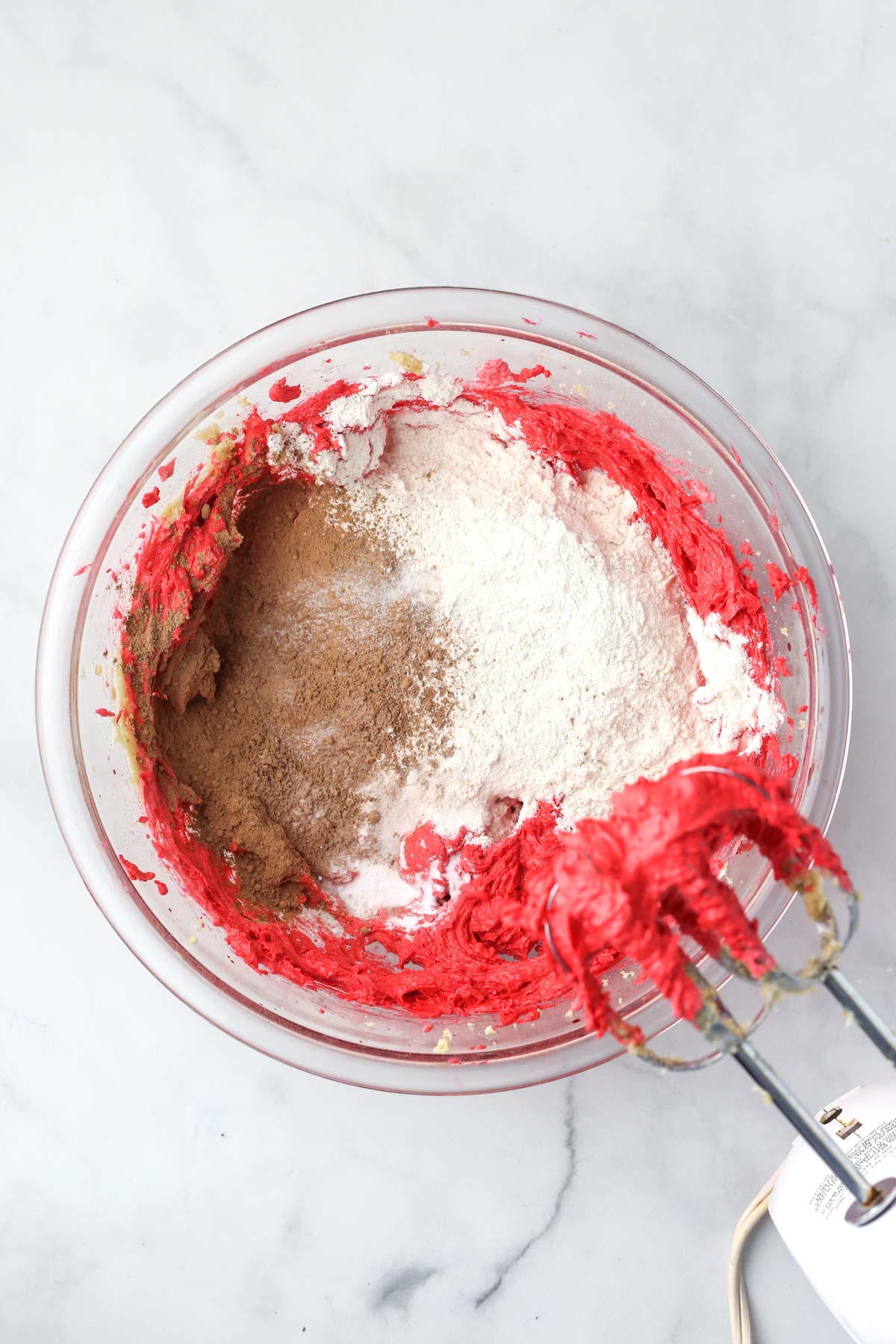 a bowl of red cookie dough with flour and cacao powder on top, about to be mixed in.