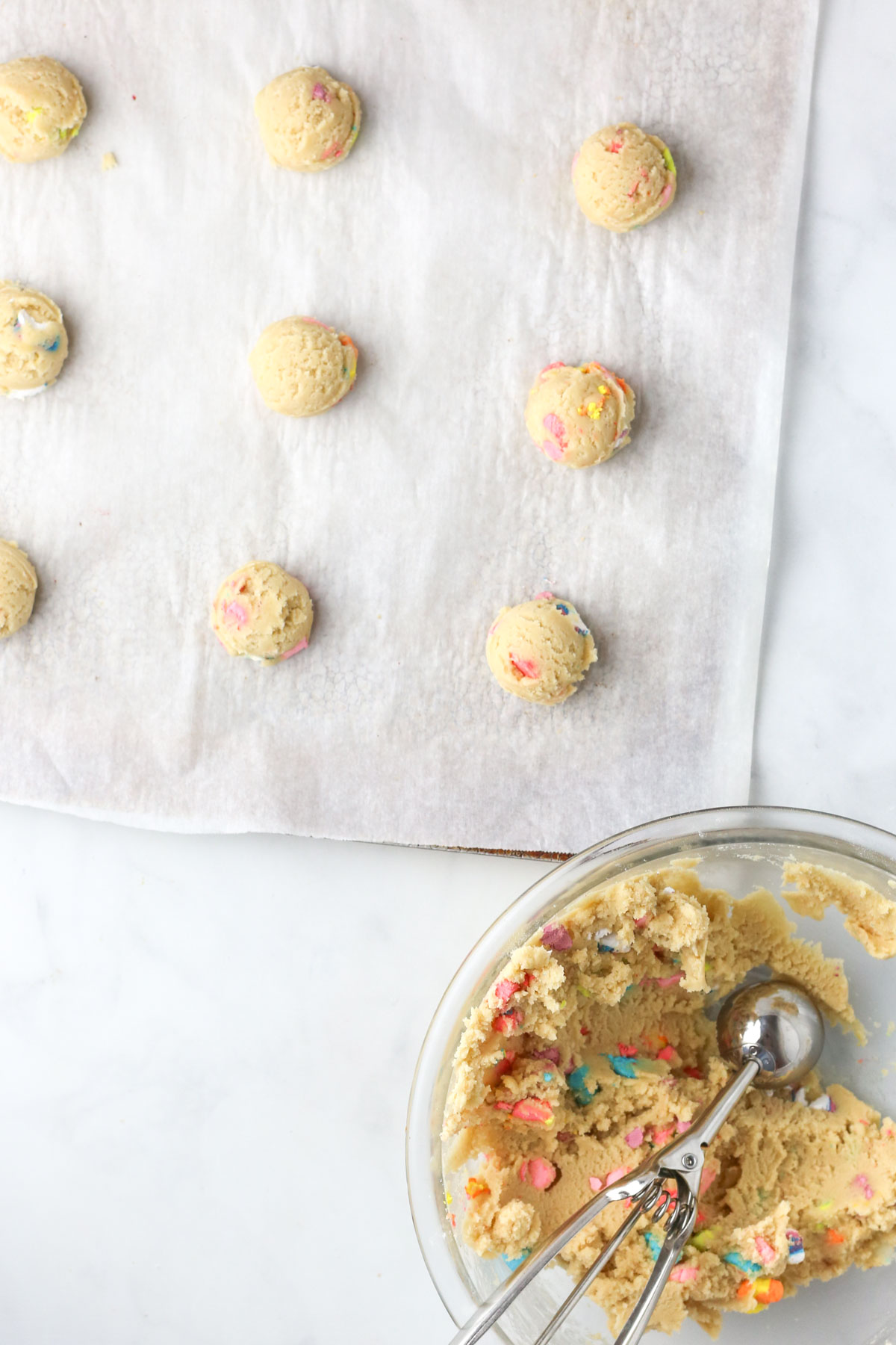 A baking tray with balls of cookie dough next to a bowl of cookie dough with a cookie scoop inside.