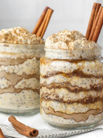 Two jars of cinnamon roll overnight oats prepared with a cinnamon swirl layer and cinnamon stick on top.