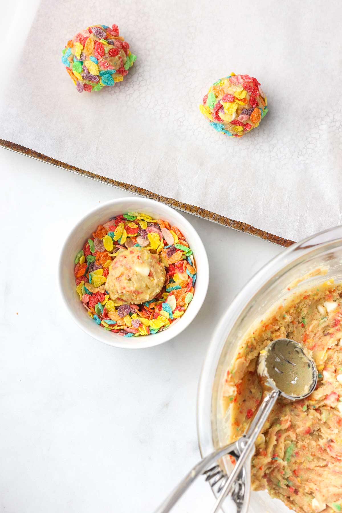 scooping cookie dough balls and rolling them in fruity pebbles cereal before placing them on the baking sheet.