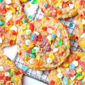 multiple freshly baked fruity pebbles cookies on a wire tray, stacked, with fruity pebbles cereal on top.