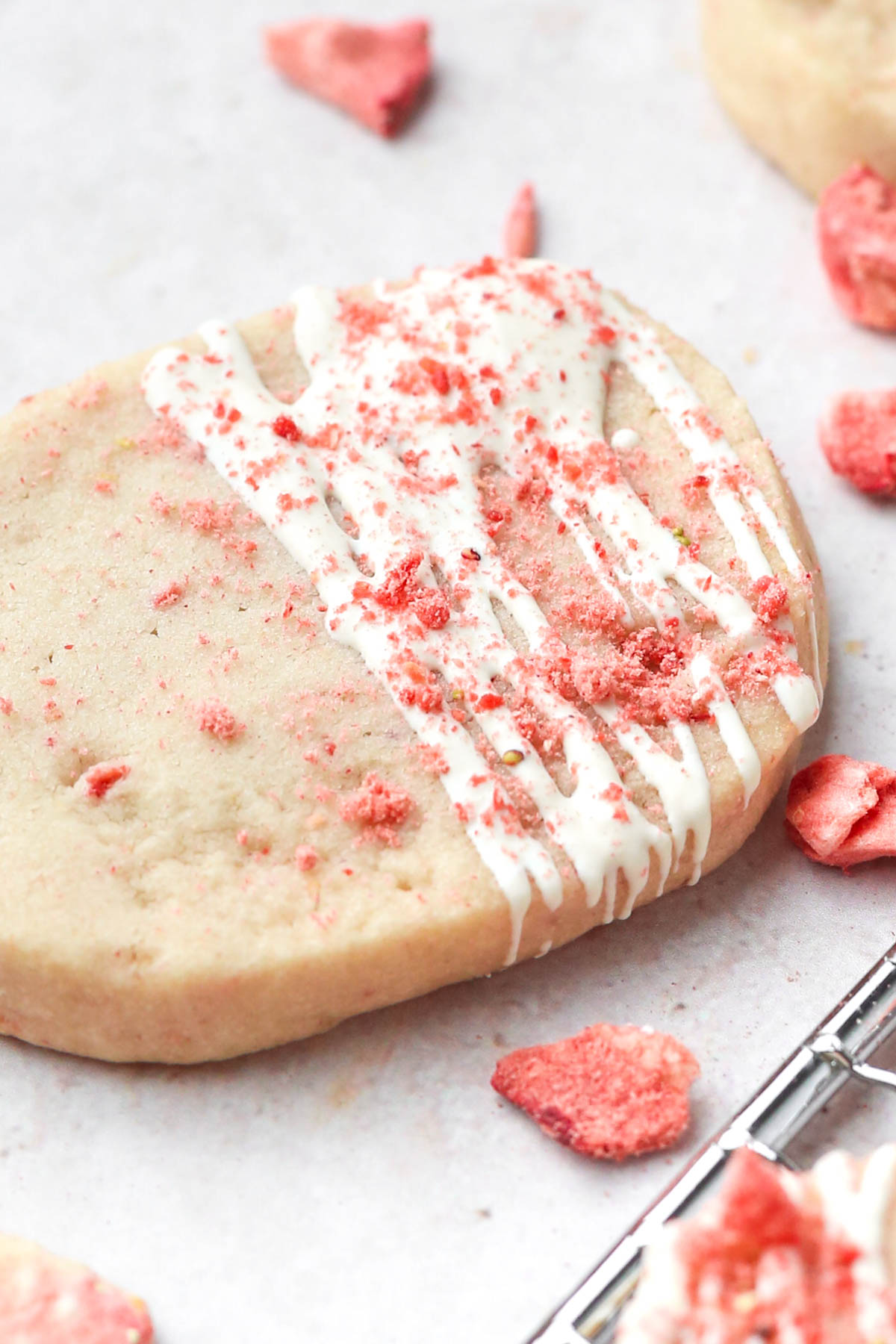 Close-up photo of a strawberry shortbread cookie with white chocolate drizzle and strawberry powder sprinkled on top, shown from an angle.