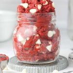 A clear jar with edible red velvet cookie dough. Inside the dough are white chocolate chips.