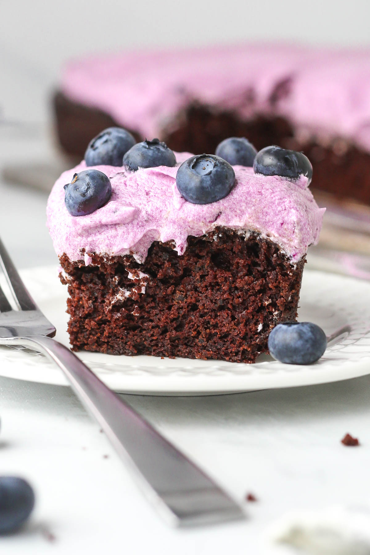 A square slice of blueberry chocolate cake with purple frosting on a small dessert plate.
